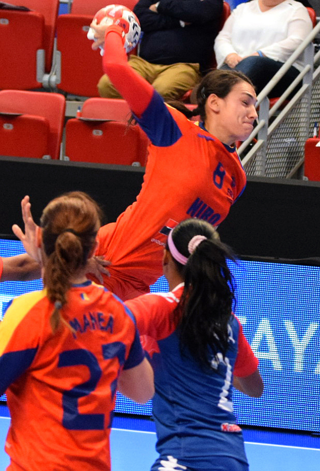 Eine Gruppe von Frauen, die in einer Halle Handball spielen, wobei eine den Ball hält und Zuschauer im Hintergrund sitzen.