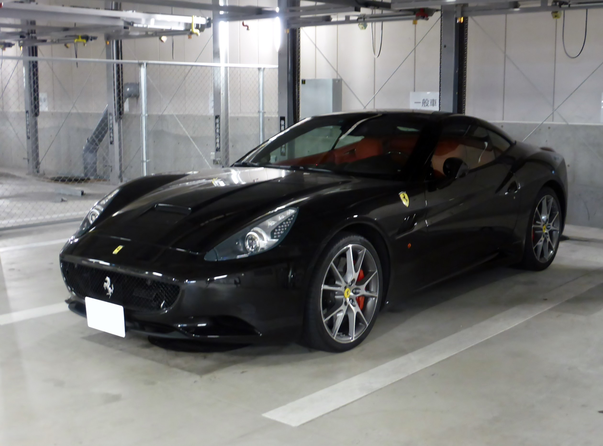 Schwarzer Ferrari California in einer Tiefgarage geparkt, mit einer Wand im Hintergrund, einem Maschendrahtzaun auf der linken Seite und Deckenlampen.
