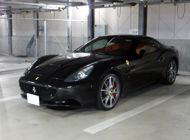 Schwarzer Ferrari California in einer Tiefgarage geparkt, mit einer Wand im Hintergrund, einem Maschendrahtzaun auf der linken Seite und Deckenlampen.