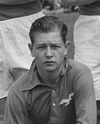 Ein Schwarz-Weiß-Foto eines Mannes auf einer Bank sitzend, der ein Hemd trägt, mit zwei Personen im Hintergrund, die angeblich der legendäre Rugbyspieler John Daly sind.