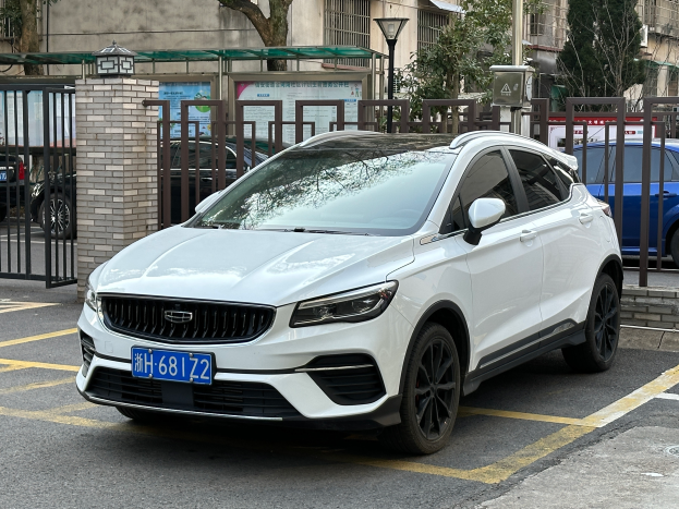Ein weißer Volvo XC40 T5 AWD Inscription auf einem Parkplatz mit Gebäuden, Bäumen, Pfählen, Lampen und anderen Gegenständen.