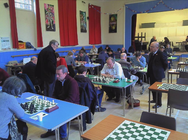 Ein großer Saal mit zwei Personen, die auf einem Tisch mit einem Schachbrett und Münzen Schach spielen, Vorhängen, Plakaten, einem Fenster und einem Kalender an der Wand.