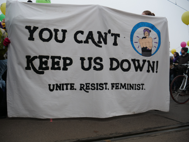 Eine Gruppe von Menschen steht auf einer Straße und hält ein Banner mit der Aufschrift "Du kannst uns nicht unterdrücken, vereine dich widerstehe Feministin", mit einem Fahrrad in der Nähe und Ballons, Drähten und einem klaren blauen Himmel im Hintergrund.