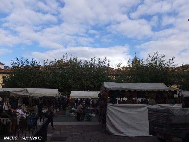 Menschen stehen und gehen in der Nähe von weißen Zelten mit Geschäften, mit Bäumen, Häusern und einem sonnigen Himmel im Hintergrund.