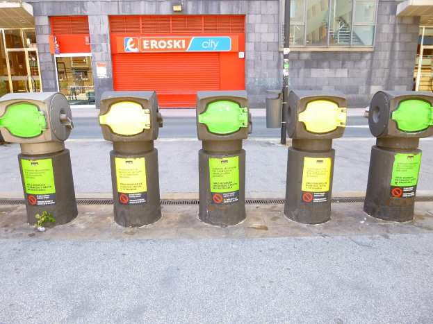 Reihe von Parkuhren entlang einer Straße mit einem Gebäude mit Glasfenstern und einem Rollladen mit Text sowie einem nahen Müllcontainer.