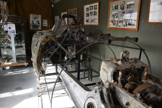 Ein altes Maschinengewehr in einem Museum ausgestellt, umgeben von Metallobjekten auf Ständern, mit einem Glaskasten links, Fotorahmen und einem Vorhang im Hintergrund.