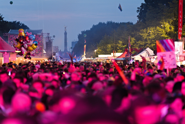 Eine große Menge bei einem nächtlichen Musikfestival, beleuchtet von bunten Lichtern, mit Zelten, Fahnen und Polen im Vordergrund und Bäumen, einem Turm und einem klaren Himmel im Hintergrund.