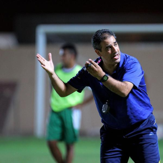 Ein Mann in einem blauen T-Shirt und Hose steht auf einem Fußballfeld und hält ein Mikrofon in der Hand, während er als Trainer gestikuliert. Im Hintergrund sind eine Person in einem grünen Kleid, ein Tor und eine Wand zu sehen.