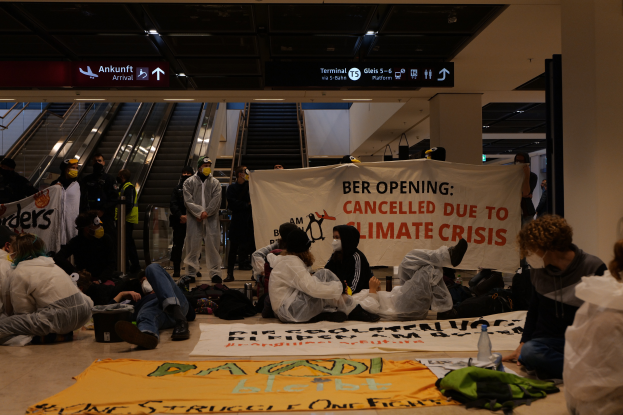 Eine Gruppe von Menschen sitzt auf einem Flughafenboden und hält ein Banner mit der Aufschrift 'Berlin wegen Klimakrise abgesagt'. Um sie herum liegen Taschen, Flaschen und andere Gegenstände verstreut, im Hintergrund sind Rolltreppen, Schilder mit Text und Deckenleuchten zu sehen.