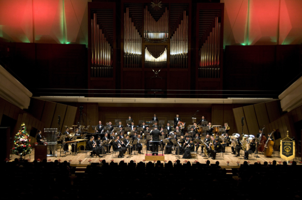 Ein großes Orchester spielt auf der Bühne vor einem Weihnachtsbaum, mit sitzenden Musikern mit Instrumenten, einem Podium mit einem Mikrofon auf der linken Seite und Texttafeln auf der rechten Seite, sowie zusätzlichen Personen unten und Beleuchtung im Hintergrund.