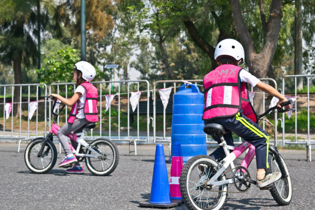 Zwei junge M├Ądchen in rosafarbenen und wei├čen Kleidern und wei├čen Helmen fahren auf Fahrr├Ądern eine Stra├če mit Verkehrshütchen entlang, mit einer blauen Trommel, einem Gel├Ąnder, B├Ąumen, einem Pfahl und Himmel im Hintergrund.