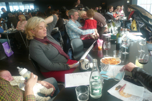 Ein Innenblick in ein belebtes Restaurant mit Menschen an Tischen, die Gläser, Teller mit Essen, Getränkeflaschen, Speisekarten und andere Gegenstände haben.