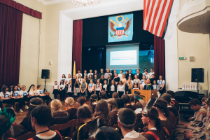 Eine Gruppe von Menschen steht an einem Podium mit einem Mikrofon während eines 2. Kunst- und Kulturwochen-Events, flankiert von Fahnen und Lautsprechern, gegenüber einer auf Stühlen sitzenden Menge, mit einem Bildschirm, der Text und ein Bild anzeigt, im Hintergrund unter einem Kronleuchter.