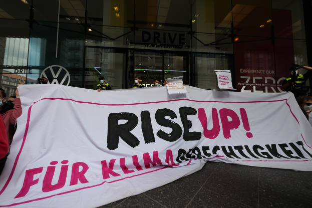 Eine Gruppe maskierter Individuals mit einem "Steh Auf"-Schild und Plakaten vor einem Glasgebäude, wahrscheinlich protestierend gegen eine deutsche Regierungsentscheidung.