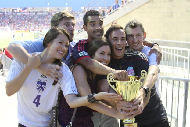 Sechs Personen, darunter zwei Frauen und vier Männer, halten einen Pokal und lächeln, posieren vor einem Eisengeländer, mit einer Wand und einem Geländer oben rechts, Menschen und Lichter im Hintergrund, möglicherweise in einem Stadion.
