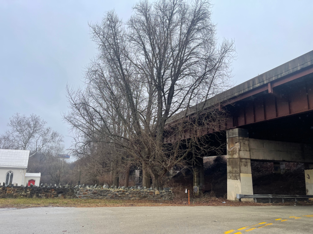 Eine Brücke über eine Straße in Reparatur, mit einem Baum in der Mitte, umgeben von verstreuten Werkzeugen, Materialien, Gras, einem Haus, einem Schild, einem Straßenschild, einer Gruppe von Bäumen und einem bewölkten Himmel.