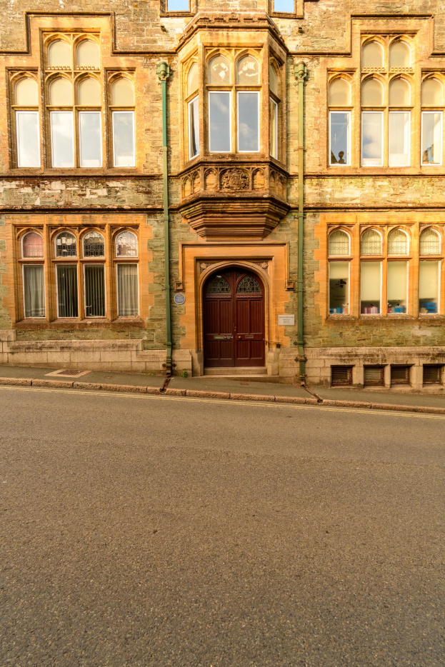 Altes Gebäude mit zahlreichen Fenstern, einer Holztür und sichtbaren Rohren an seinen Außenwänden, das neben einer Straße steht.