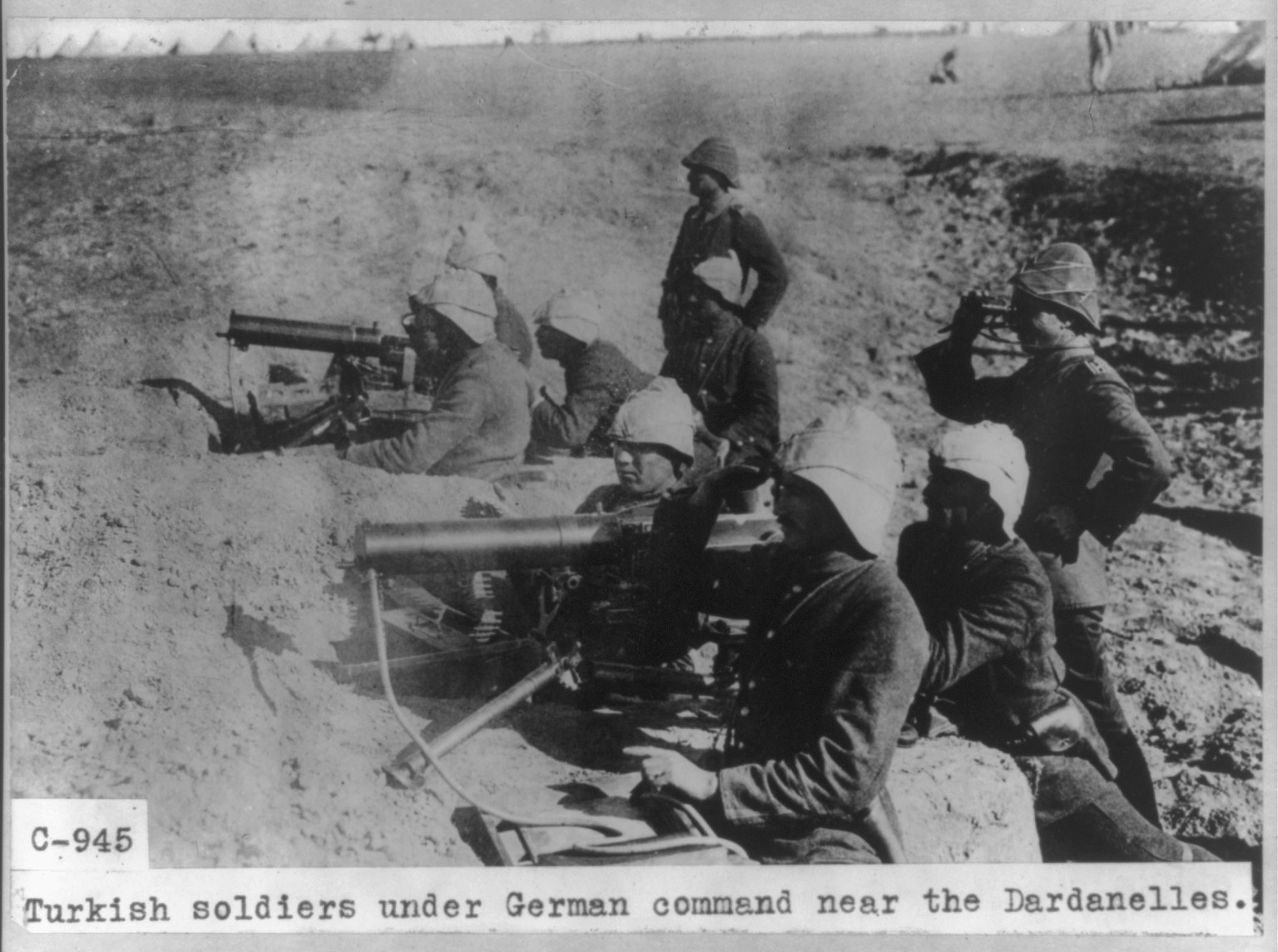 Schwarzes und weißes Foto von türkischen Soldaten unter deutschem Kommando in der Nähe der Dardanellen, die Helme tragen und Waffen halten, mit Text unten.
