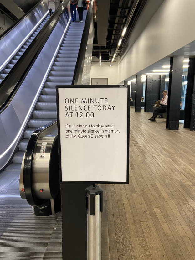 Eine Rolltreppe im Flughafen mit einem Schild, auf dem "Eine Minute Schweigen heute" steht, einige Menschen darauf und Lampen an der Decke im Hintergrund.