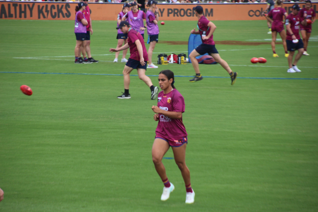 Frauen beim Australian-Rules-Fußball auf einem grünen Feld mit verstreuten Bällen und einem Banner im Hintergrund, einige tragen Kappen und Turnschuhe.