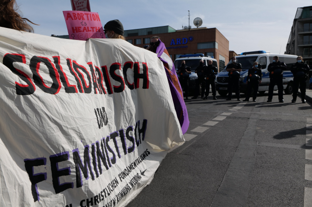 Eine Gruppe von Menschen marschiert die Straße entlang und hält ein Banner mit der Aufschrift 'Solidarität und Feminismus', mit parkenden Fahrzeugen und Gebäuden im Hintergrund unter einem klaren blauen Himmel.