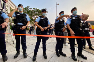 Eine Gruppe von Polizisten in Masken und Mützen steht vor einem Gebäude, mit einer Schleife im Vordergrund und einer Person mit einer Kamera auf der linken Seite, vor einem Hintergrund aus Bäumen, Laternenmasten, Gebäuden und einem klaren blauen Himmel.