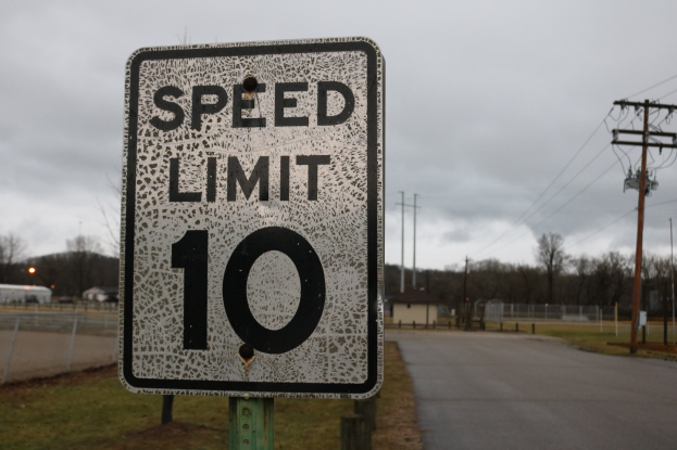 Geschwindigkeitsbegrenzungstafel am Rande einer Straße mit Gras, Strommästen, einem Zaun, Häusern, Bäumen und einem bewölkten Himmel im Hintergrund.