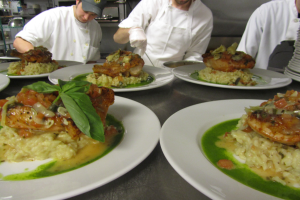 Drei Köche in einer Küche, die Teller mit Speisen vorbereiten, tragen Mützen und Handschuhe, mit Regalen im Hintergrund.