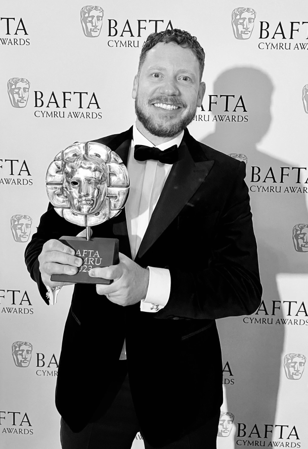 Ein Mann in einem Smoking, der eine BAFTA-Trophäe hält und lächelt, mit einem Hintergrundplakat, auf dem "BAFTA Awards 2018" steht.