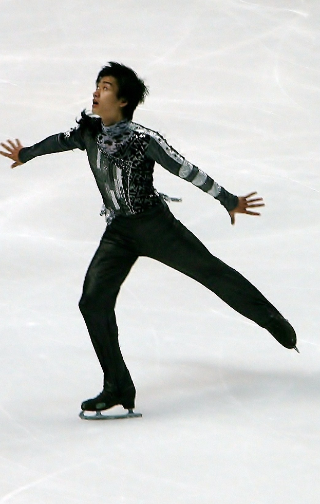 Männlicher Eiskunstläufer in einem schwarzen Kleid, der eine Choreographie auf einem Eisstadion präsentiert