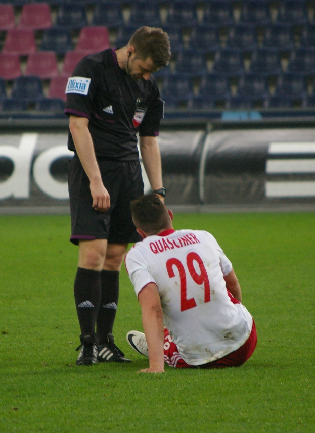 Ein Füßballspieler sitzt neben einem Schiedsrichter auf dem Boden, beide in Sportkleidung, mit Stadiontafeln und Stühlen im Hintergrund.