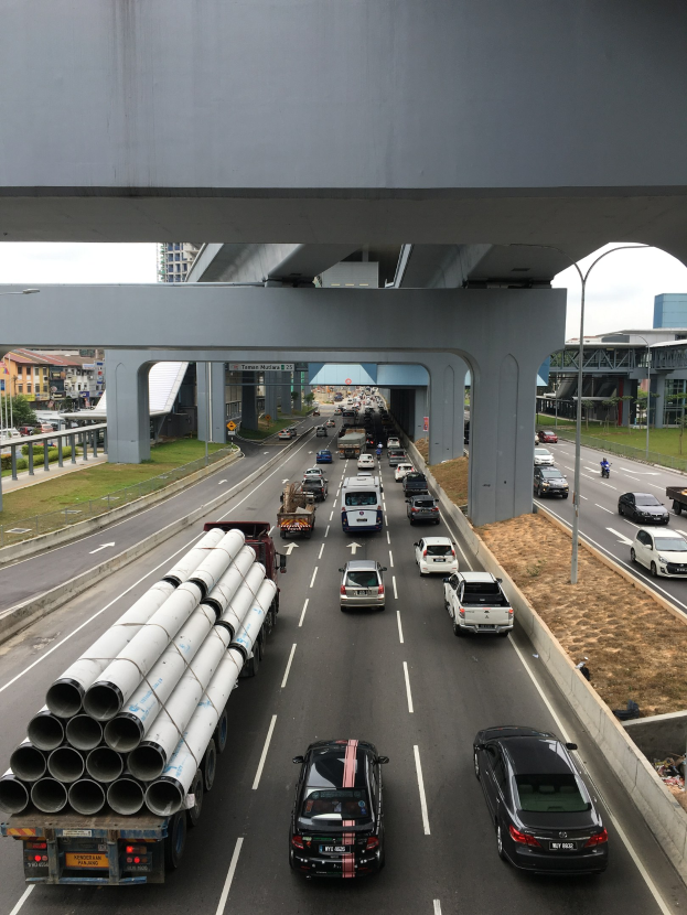 Eine vielbefahrene Autobahn mit zahlreichen Fahrzeugen, Verkehr unter einer Brücke, Straßenlaternen, Gras, Gebäuden, Bäumen und dem Himmel.