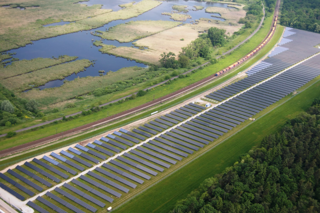 Luftaufnahme einer Solaranlage mit Panelen in einem Feld, umgeben von Bäumen, Gras, Pflanzen und Wasser, mit einem Zug auf einer nahen Eisenbahnschiene.