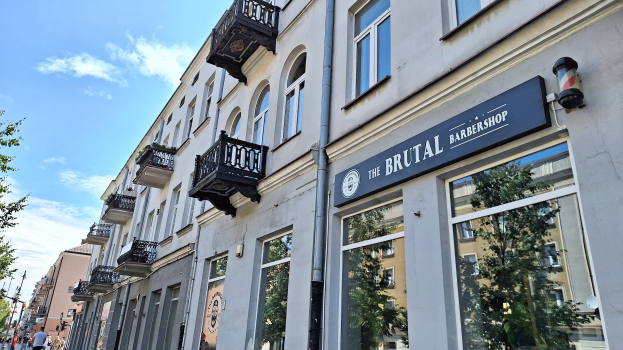 Brutalistischer Barbershop in Berlin mit eckigen Betongebäuden, Fenstern, Geländern, Balkonen und Namensschildern, umgeben von Bäumen, Straßenlaternen und Fußgängern unter einem bewölkten Himmel.