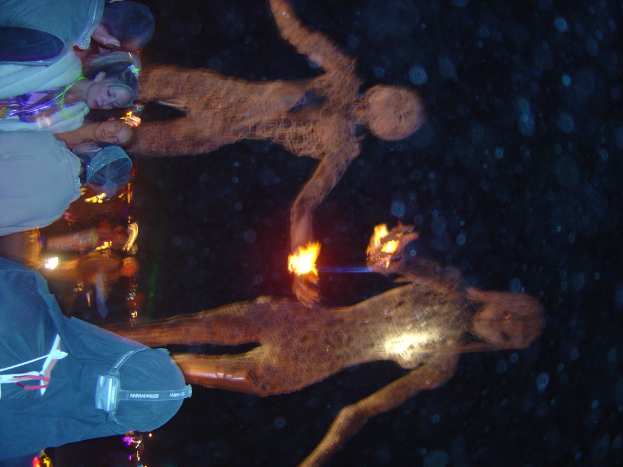 Eine Gruppe von Menschen auf der linken Seite des Bildes mit brennenden Statuen dahinter und Lichtern im Hintergrund.