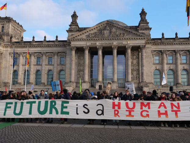 Eine Gruppe von Menschen hält ein Transparent mit der Aufschrift "Zukunft ist ein Menschenrecht" vor dem Reichstagsgebäude in Berlin, Deutschland, das mit Fenstern, Säulen und Bögen verziert ist und von Fahnenmasten umgeben ist.