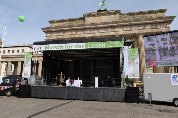 Eine Bühne vor dem Brandenburger Tor in Berlin, Deutschland, mit einem Tisch, einer Vase mit Blumen, einem Kreuzsymbol, Fahnen, Lautsprechern und anderen Gegenständen, sowie Fahrzeugen auf der Straße und Gebäuden im Hintergrund.