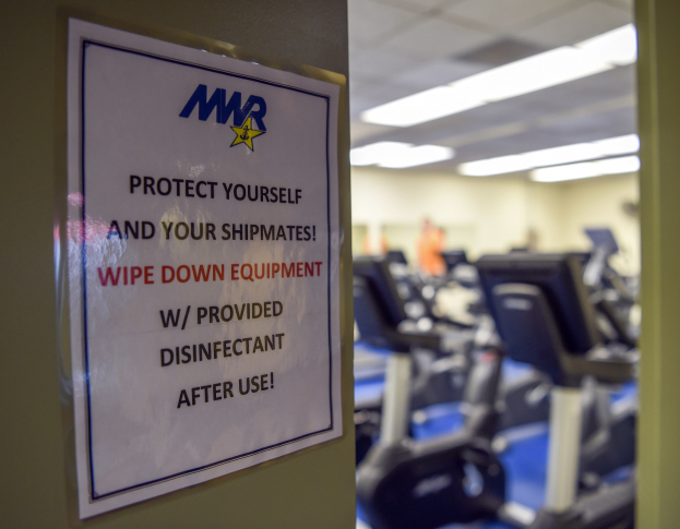 Ein Schild an einer Gym-Tür mit der Aufschrift "Schützen Sie sich und Ihre Schiffskameraden" mit Laufbändern und Deckenleuchten im Hintergrund, das Bild leicht unscharf, um Bewegung zu suggerieren.