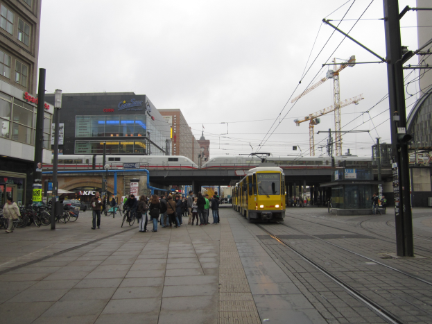 Eine gelbe Straßenbahn auf einer Stadtstraße mit Menschen und Fahrrädern auf dem Gehweg, Pfosten, Drähte, eine Brücke, ein Zug, Gebäude, Schilder, ein Kran und ein bewölkter Himmel.