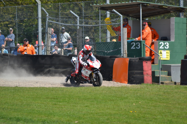 Eine Person in Helm und Handschuhen fährt ein Motorrad über ein grasbewachsenes Feld, mit Zuschauern in Kappen und Schutzbrillen, einem eingezäunten Bereich, Treppen mit Geländern und einem nummerierten Schuppen im Hintergrund.