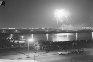 Ein Schwarz-Weiß-Foto einer Menge, die ein Feuerwerk über Wasser betrachtet, mit Fahrzeugen, Laternenpfählen, Bäumen und Gebäuden im Hintergrund.