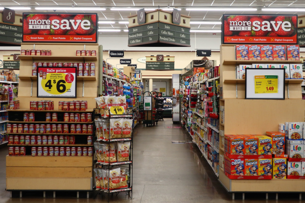 Ein Gang eines Lebensmittelgeschäfts mit verschiedenen Lebensmitteln auf Regalen und dem Boden, mit beleuchteter Decke und Informationsschildern im Hintergrund und einer Person weiter hinten.