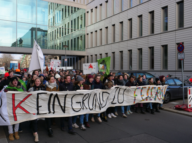 Eine Gruppe von Menschen steht auf der Straße und hält ein Schild mit der Aufschrift "Kein Grind Lo Feuer" hoch, mit Gebäuden, Bäumen, Polen, Schildern, Fahrzeugen und einem klaren blauen Himmel im Hintergrund.