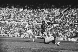 Schwarzes Foto von Männern beim Fußballspielen auf einem Feld mit Zuschauern im Hintergrund und einer 'Bundesliga 1978-79 Ajax 1-1 Hannover 96'-Fahne.