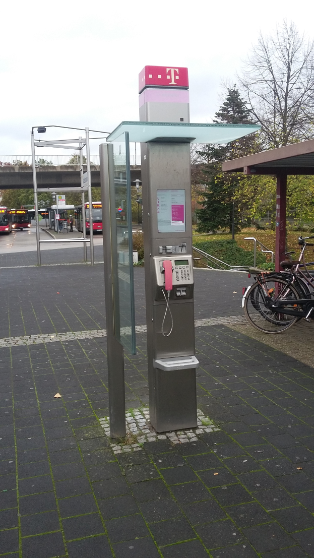 Ein Münzfernsprecher steht an der Straße neben einer Bushaltestelle, daneben parkt ein Fahrrad, während im Hintergrund Bäume, Fahrzeuge, eine Brücke und ein klarer blauer Himmel zu sehen sind.