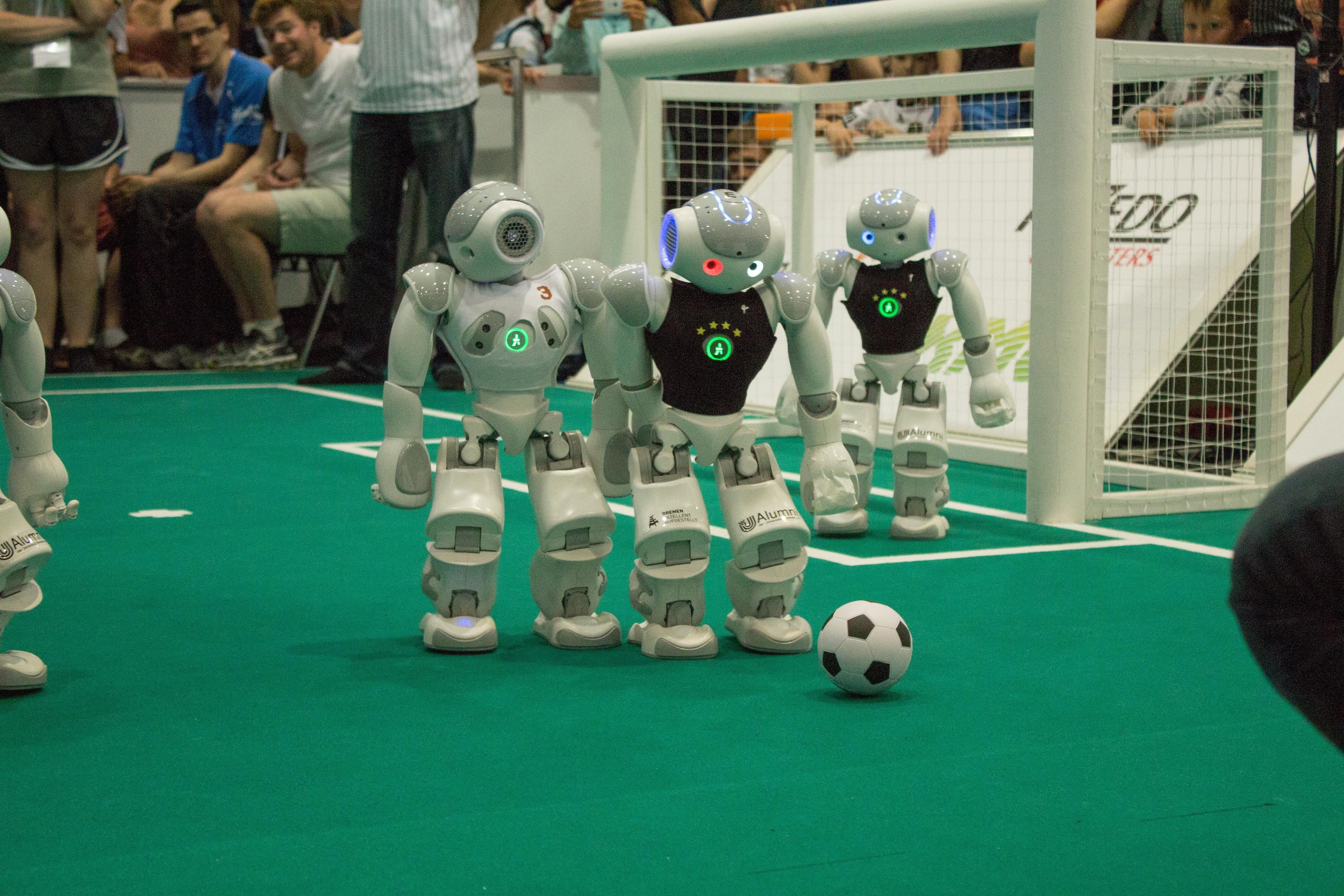 Eine Gruppe von Robotern, die auf einem grünen Feld Fußball spielen, mit einem Ball in der Mitte, einem Netz auf der rechten Seite und Zuschauern im Hintergrund, die Kameras halten.