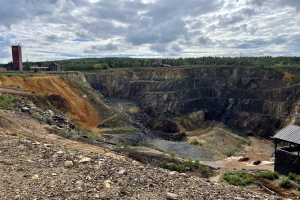 Großer Tagebau auf einem Schotterfeld umgeben von Bäumen, Gebäuden und einem Schuppen unter einem bewölkten Himmel, mit verstreuten Steinen und Pflanzen.