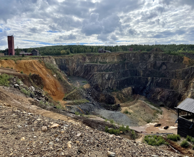 Großer Tagebau auf einem Schotterfeld umgeben von Bäumen, Gebäuden und einem Schuppen unter einem bewölkten Himmel, mit verstreuten Steinen und Pflanzen.