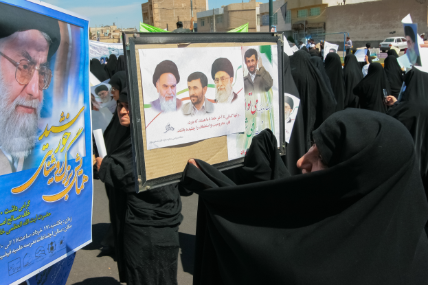 Eine Gruppe von Frauen in schwarzen Abayas hält Plakate mit Fotos von Ajatollah Ali Khamenei hoch, mit Gebäuden, Polen, Fahrzeugen und einem klaren blauen Himmel im Hintergrund.