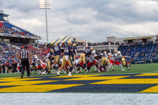 Ein Fussballspiel im Gange mit Spielern in Sportbekleidung und Helmen im Vordergrund, Zuschauern, Geländern, Tafeln mit Text, Lichtmasten und einem bewölkten Himmel im Hintergrund und Text unten auf dem Bild.